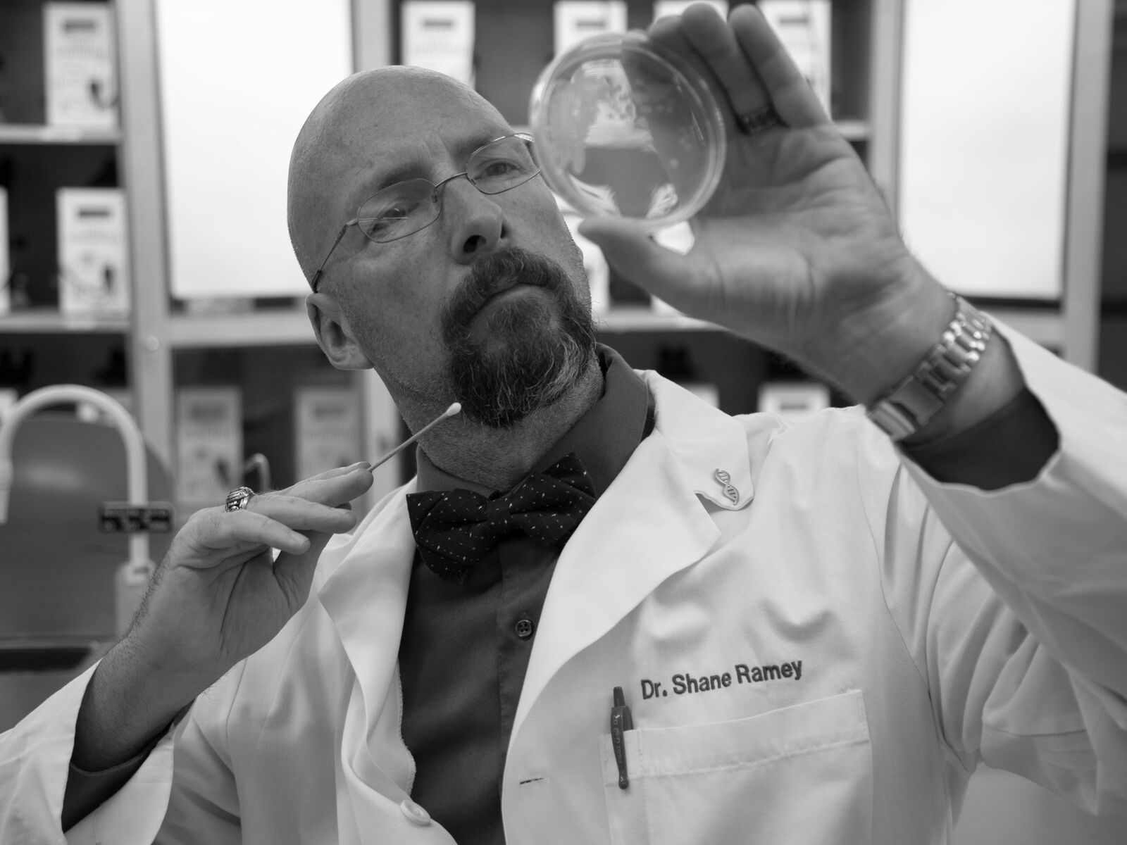 Dr. Ramey wearing a lab coat and bowtie, holding a Q-Tip and peering into a glass dish. 