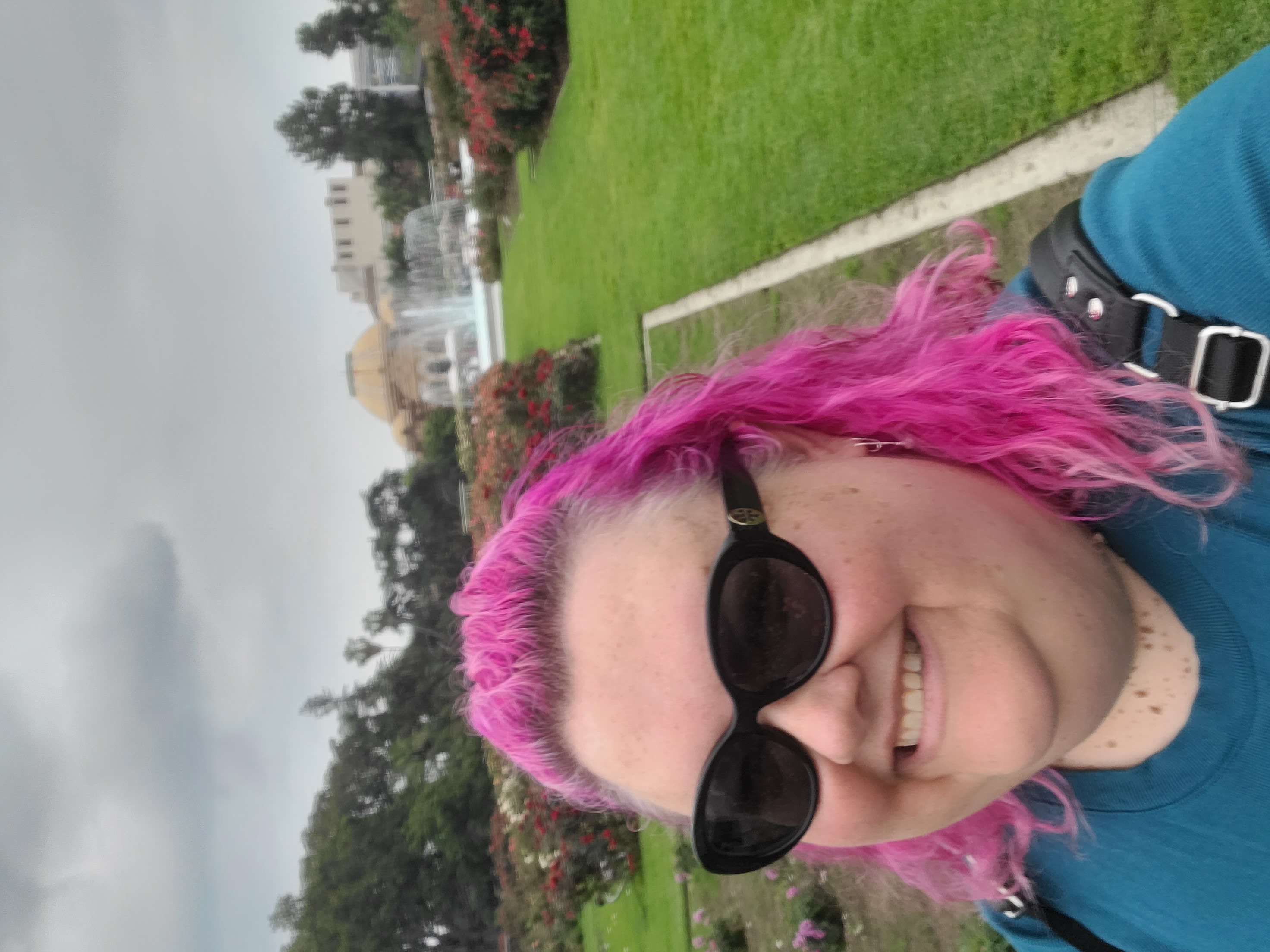 A woman with pink hair, sunglasses, and a blue shirt smiles with a fountain and the Natural History Museum in the background