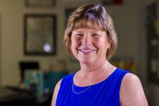 A photo of the instructor with a blurred background of a classroom. The instructor is smiling at the camera. She has short light brown hair with bangs and is wearing a blue sleeveless dress.
